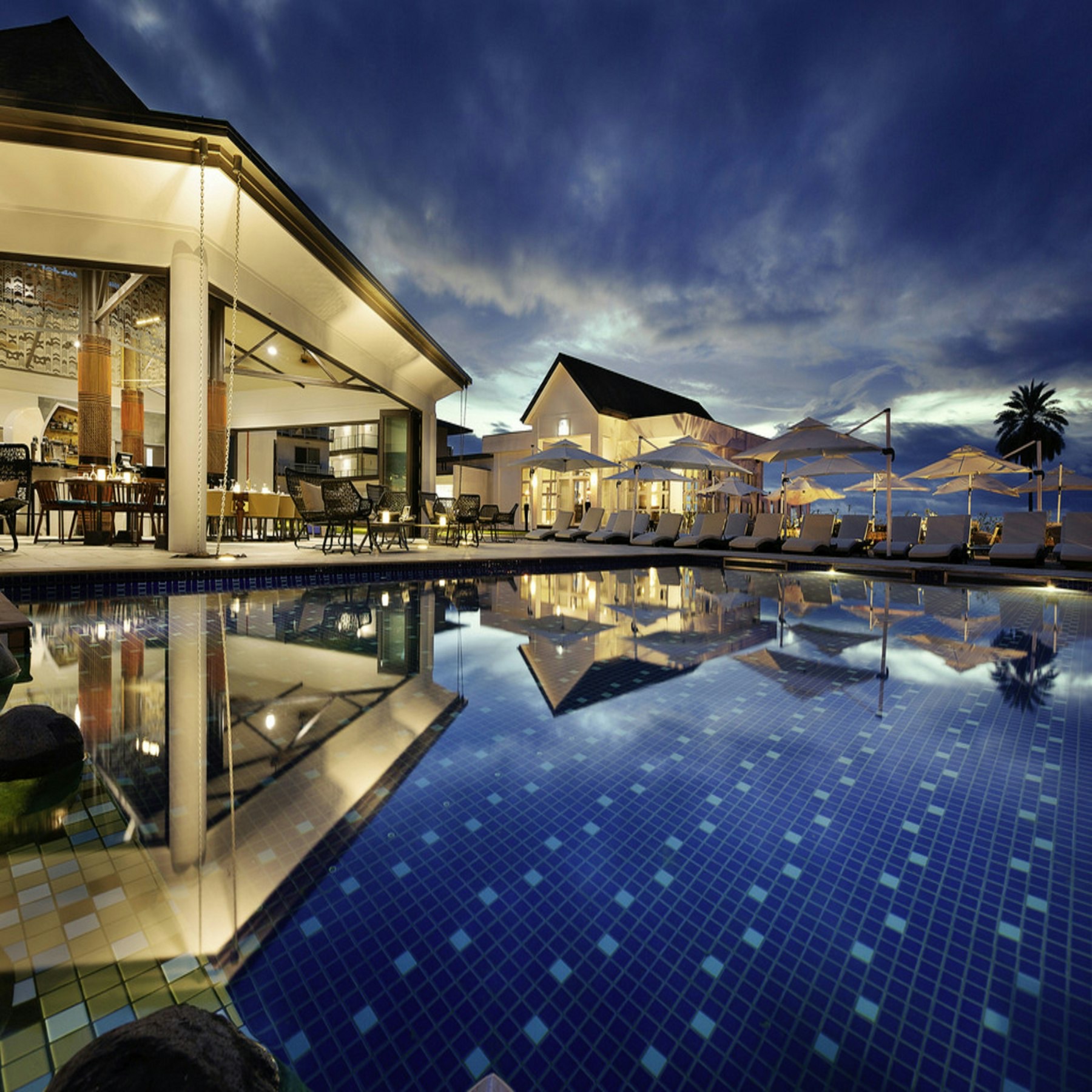 A resort pool at dusk, with lounge chairs and a lit dining terrace